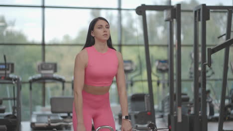A-hispanic-brunette-woman-in-a-pink-suit-pulls-the-barbell-to-the-top-while-training-her-shoulders-in-the-gym.-Standing-exercise-for-training-the-shoulders-and-arms.-Weight-training